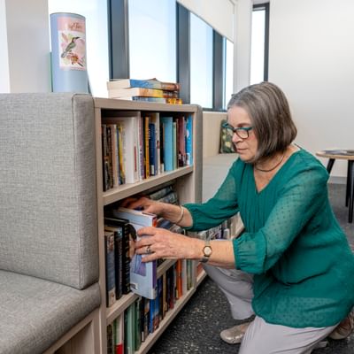 A person with short gray hair in a teal blouse kneels beside a bookshelf, selecting a book.