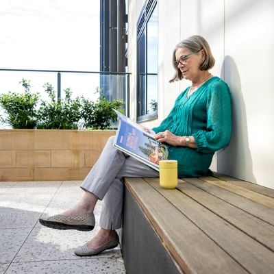 Older person in teal blouse sits on a wooden bench reading a magazine; yellow mug beside, greenery behind.
