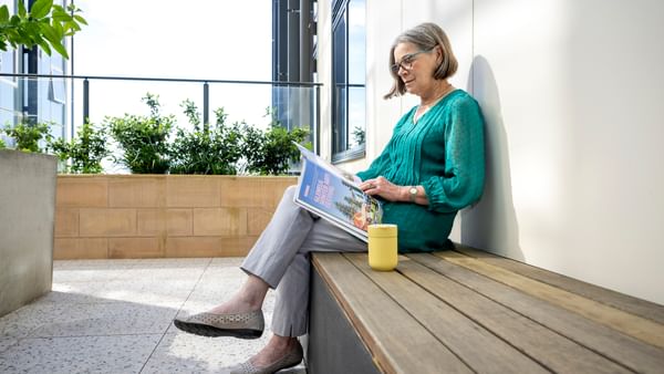 Older person in teal blouse sits on a wooden bench reading a magazine; yellow mug beside, greenery behind.