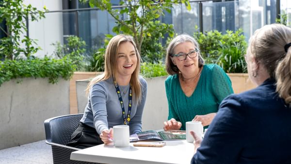 Three people sit at an outdoor table with mugs and a brochure, smiling.