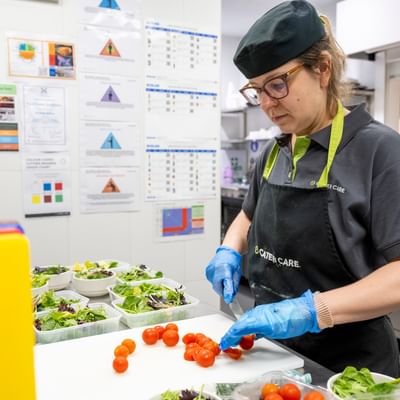 Cater Care staff member wearing cap and apron chops cherry tomatoes on a cutting board in a kitchen.