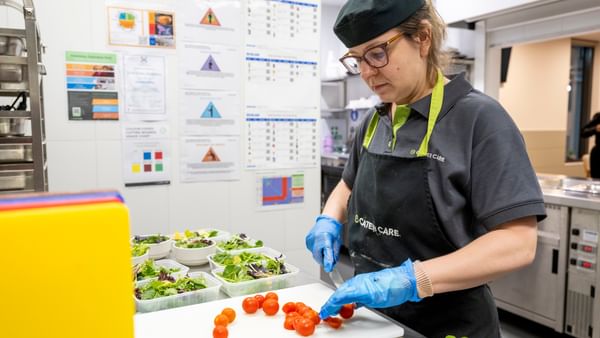 Cater Care staff member wearing cap and apron chops cherry tomatoes on a cutting board in a kitchen.