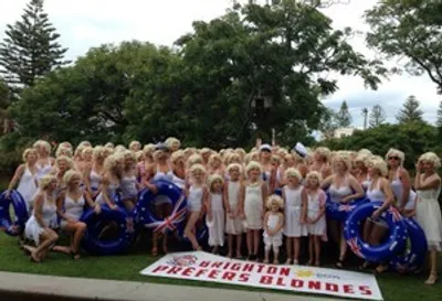 Group of people in white and blue dresses outdoors with a banner that reads Brighton Prefers Blondes