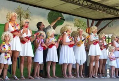 Group of performers in white dresses with leis on a stage, tropical backdrop.