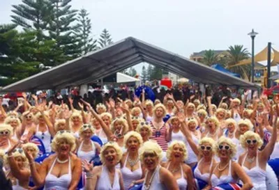Large crowd wearing white outfits and blonde wigs, raising arms under a canopy at an outdoor event.