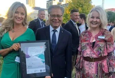 Outdoor event; a person in a suit stands between two others, one of whom holds a framed certificate.