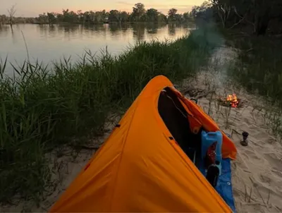 Orange tent on a sandy riverbank with tall grass, a small campfire, and camping gear nearby.