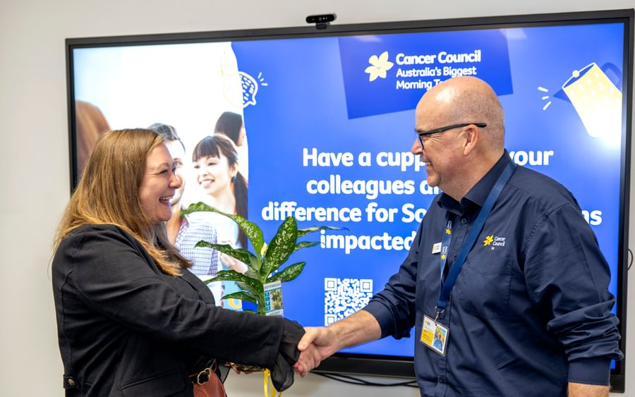 A woman and a man shake hands in front of a Cancer Council SA screen