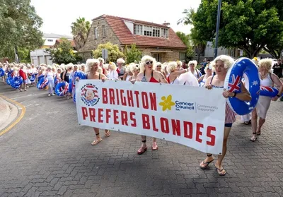 People wearing blonde wigs march in a parade, carrying a banner reading Brighton Prefers Blondes.