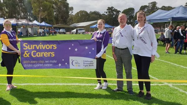 Four people on a grassy field hold a purple 'Survivors & Carers' banner at a cancer charity event.