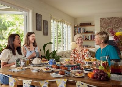 Four people chat around a sunlit table filled with cakes and fruit, in a bright room with plants.