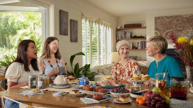 Four people chat around a sunlit table filled with cakes and fruit, in a bright room with plants.