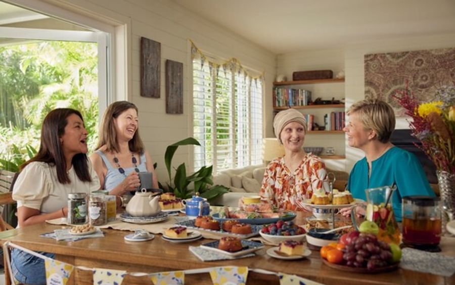 Four people chat around a sunlit table filled with cakes and fruit, in a bright room with plants.