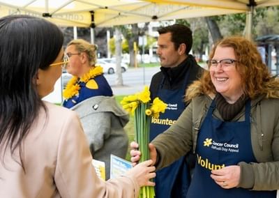 Smiling volunteer hands yellow daffodils to a guest under a canopy at an outdoor event.