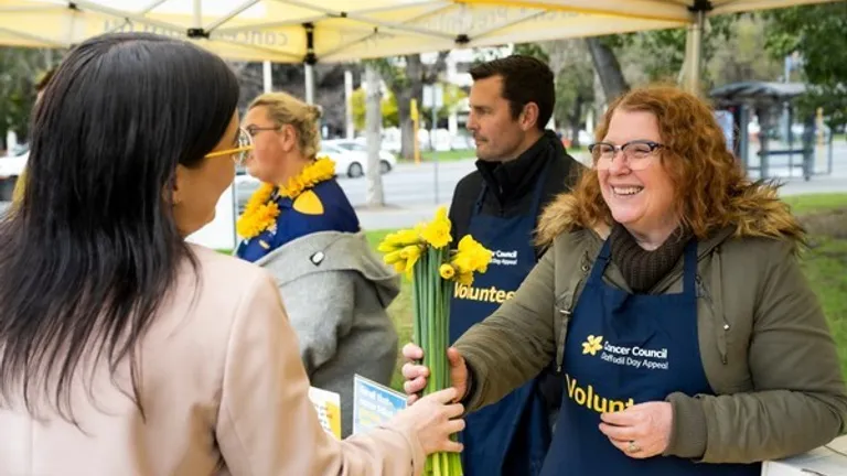 Smiling volunteer hands yellow daffodils to a guest under a canopy at an outdoor event.
