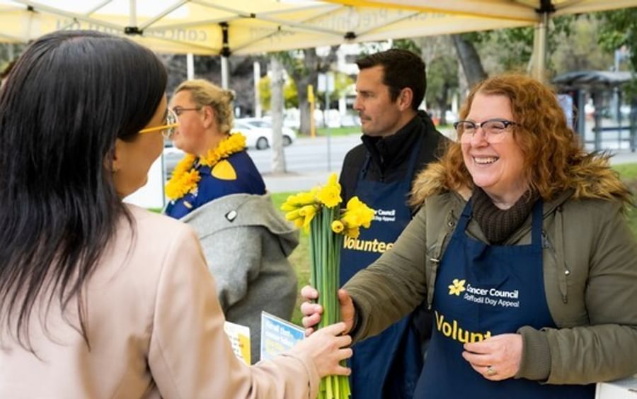 Smiling volunteer hands yellow daffodils to a guest under a canopy at an outdoor event.