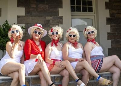 Five people in retro white outfits with wigs, red accessories and sunglasses sit on a verandah.
