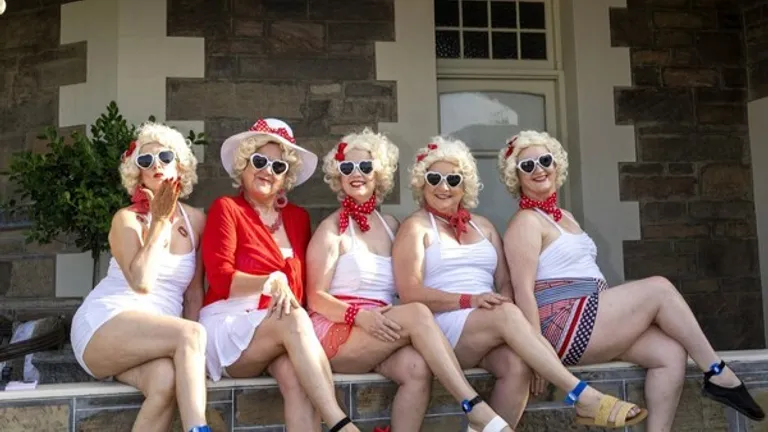 Five people in retro white outfits with wigs, red accessories and sunglasses sit on a verandah.