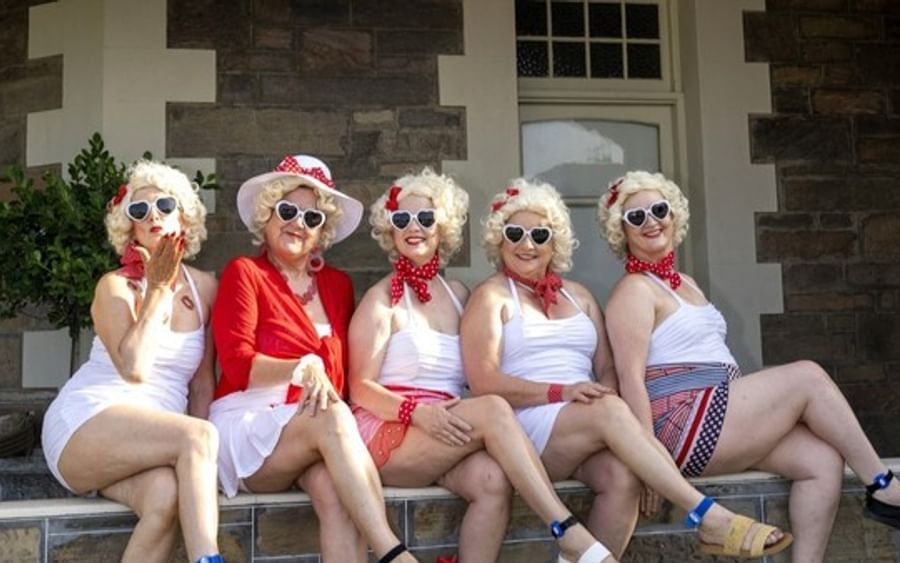 Five people in retro white outfits with wigs, red accessories and sunglasses sit on a verandah.
