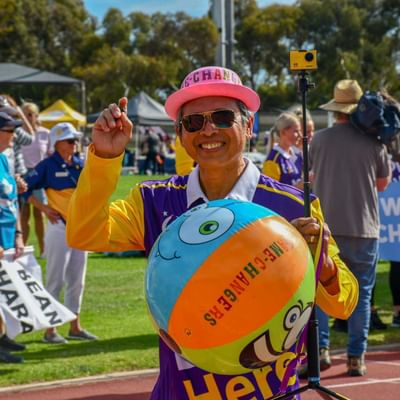 Smiling person in yellow and purple holds a beach ball on a selfie stick at a charity walk.