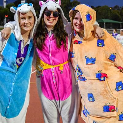 Three people in colourful onesies pose with arms around each other on a track field, smiling.