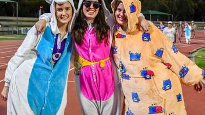 Three people in colourful onesies pose with arms around each other on a track field, smiling.