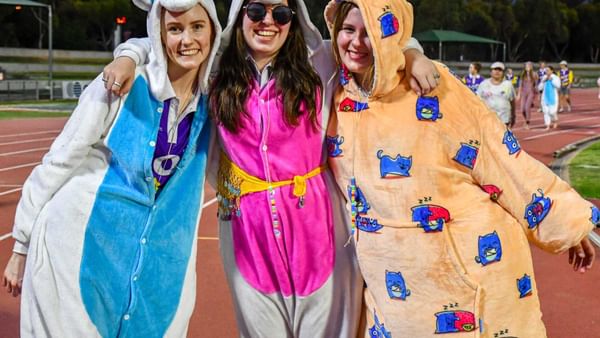 Three people in colourful onesies pose with arms around each other on a track field, smiling.