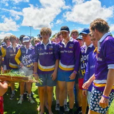 Group of youths in purple Relay For Life shirts on a sunny field as someone holds a basket of grass.