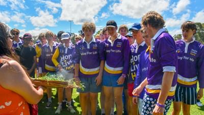 Group of youths in purple Relay For Life shirts on a sunny field as someone holds a basket of grass.