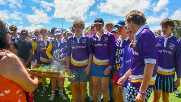 Group of youths in purple Relay For Life shirts on a sunny field as someone holds a basket of grass.