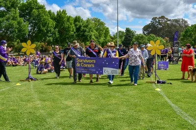 Group of people walk across a sunny park carrying a purple Survivors & Carers banner, flanked by yellow flower props.