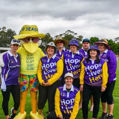 Group of people in purple Hope Lives Here shirts posing with a yellow seagull mascot on a field.