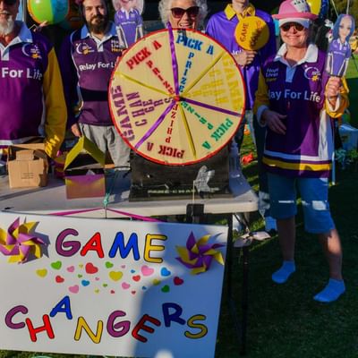 Group of people in purple Relay For Life shirts around a prize wheel on a field, GAME CHANGERS sign.