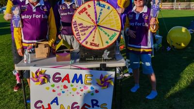 Group of people in purple Relay For Life shirts around a prize wheel on a field, GAME CHANGERS sign.