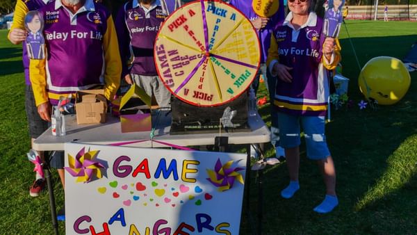 Group of people in purple Relay For Life shirts around a prize wheel on a field, GAME CHANGERS sign.