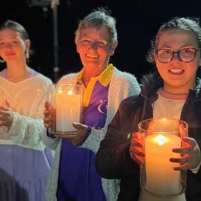 Three people at night hold large lit candles in glass jars, smiling at the camera.