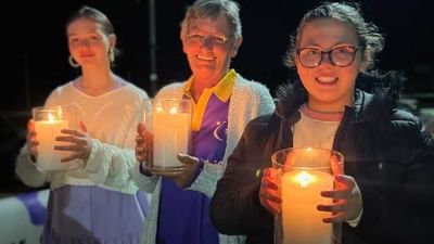 Three people at night hold large lit candles in glass jars, smiling at the camera.