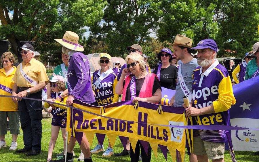 Group of people in purple and yellow hold an Adelaide Hills banner at a Relay For Life event.