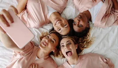 Four people lying on a bed in pink robes, taking a selfie with a pink mobile and smiling.