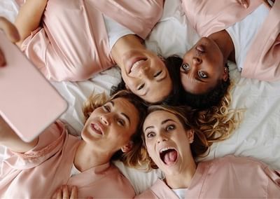 Four people lying on a bed in pink robes, taking a selfie with a pink mobile and smiling.