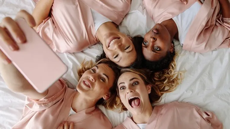 Four people lying on a bed in pink robes, taking a selfie with a pink mobile and smiling.