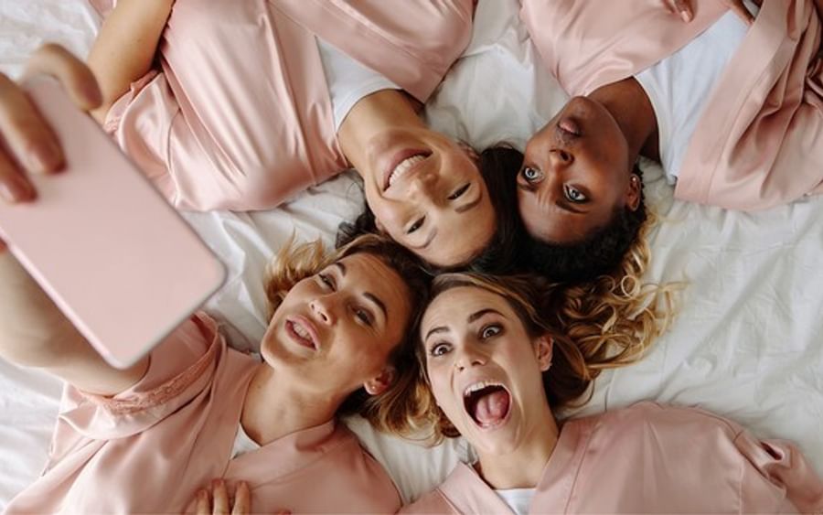 Four people lying on a bed in pink robes, taking a selfie with a pink mobile and smiling.