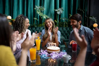 Group of friends clapping and smiling around a birthday cake with candles at a table.