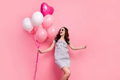 Person in a white polka-dot outfit, smiling, holding pink balloons against a pink backdrop.