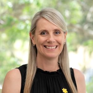 Smiling person with blonde hair, wearing a black sleeveless top and a yellow flower pin, outdoors.