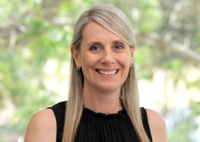 Smiling person with blonde hair, wearing a black sleeveless top and a yellow flower pin, outdoors.