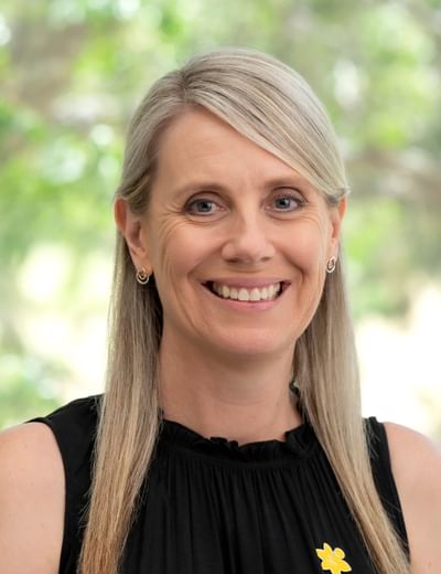 Smiling person with blonde hair, wearing a black sleeveless top and a yellow flower pin, outdoors.