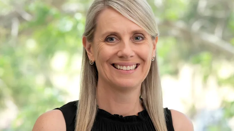 Smiling person with blonde hair, wearing a black sleeveless top and a yellow flower pin, outdoors.