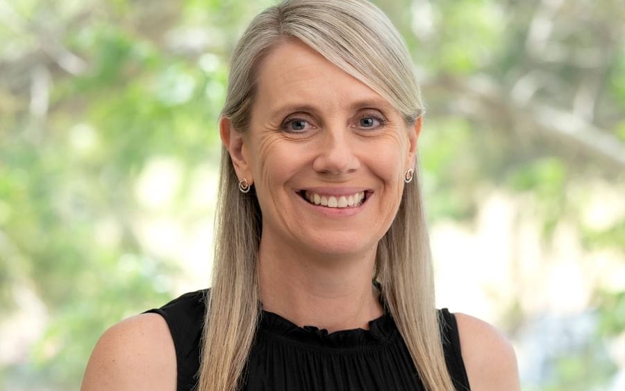 Smiling person with blonde hair, wearing a black sleeveless top and a yellow flower pin, outdoors.