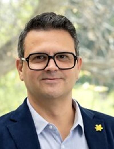 Portrait of a person in a blue blazer and light shirt, wearing glasses, outdoors.